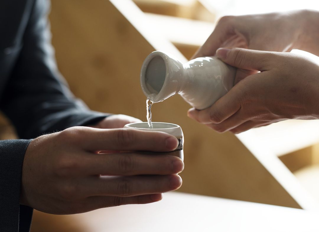 Person pouring liquid from a small white container into a cup held by another person.